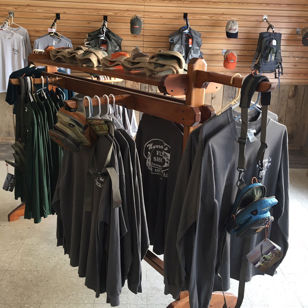 Shirts, hats and fishing vests and fishing packs shown on a wooden rack inside our fly shop