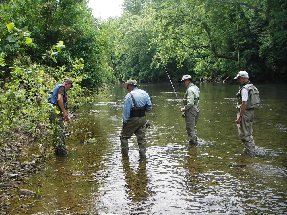 Three student anglers learning about minnows and where they are found in the river from Dale, the instructor all four are standing in a clear river carrying fly rods and wearing fishing waders and vests 