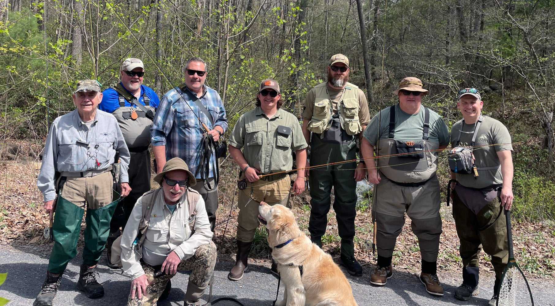 Fishing class group photo with anglers standing on the gravel road in a group many of which are holding fly rods surrounded by a beautiful forest with the trout stream in the background