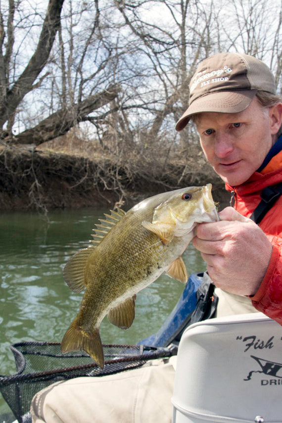 An angler holding a nice smallmouth bass caught on a crayfish.  The angler is sitting in a seat on a boat in the late winter with a landing net in the background and trees along the riverbank are visible without leaves on them.