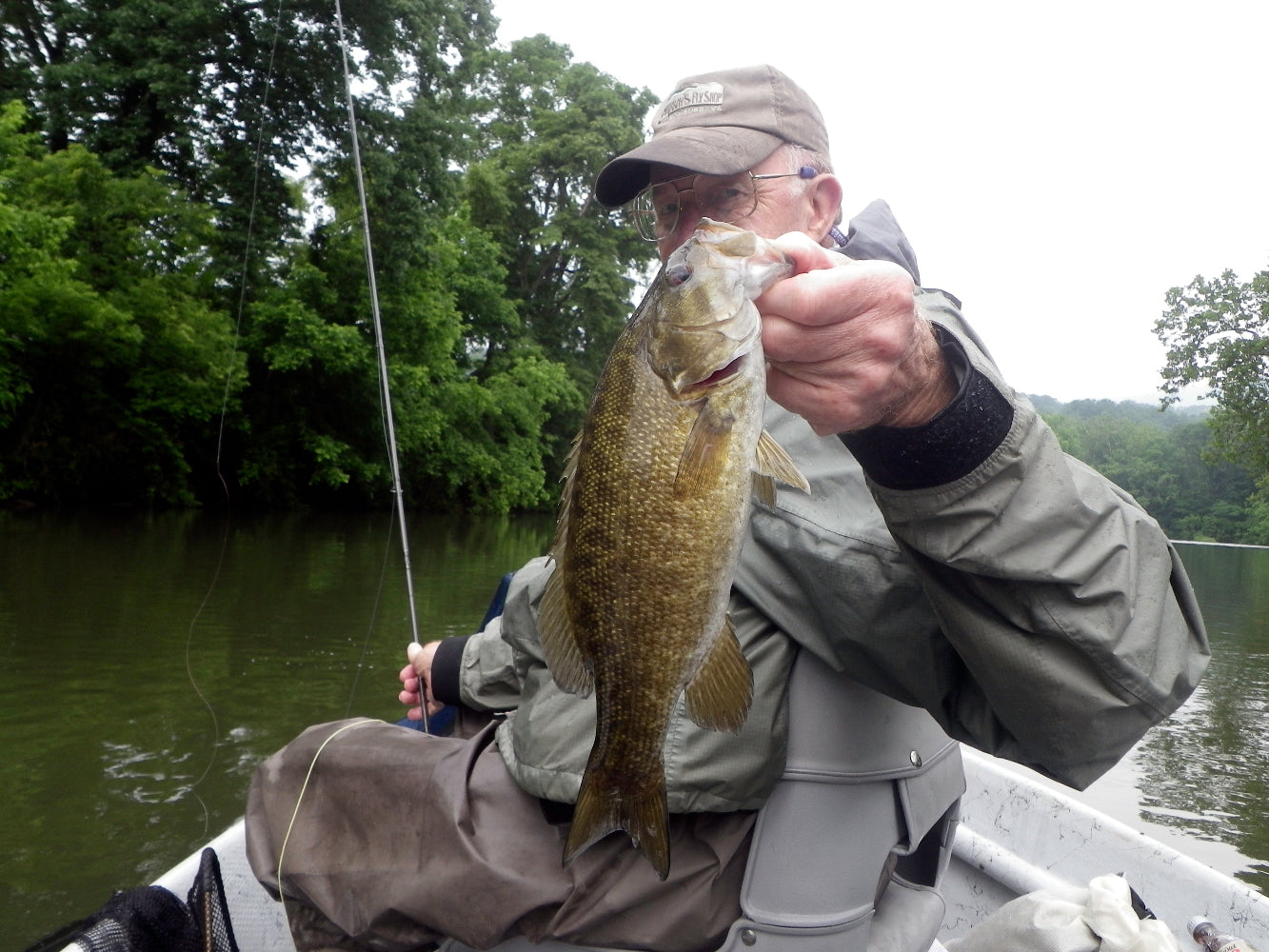 Harry Murray holding a nice smallmouth bass he caught on a fly rod in a Hyde drift boat on the Shenandoah River in Virginia.  He is wearing a raincoat and fishing waders, holding a fly rod surrounded by water and green trees along the river bank